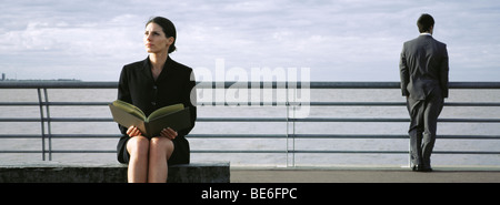 Businesswoman on bench at waterfront avec livre ouvert sur les genoux, l'homme au garde-fous à la vue à en arrière-plan Banque D'Images