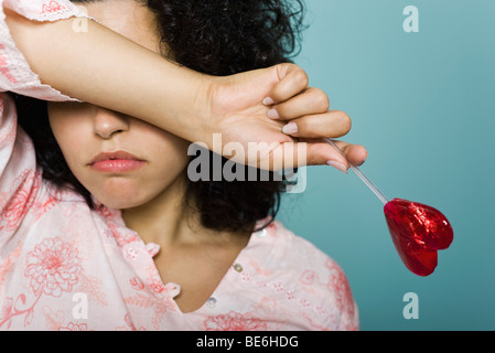 Femme froncer et couvrant les yeux avec le bras, holding heart-shaped lollipop Banque D'Images