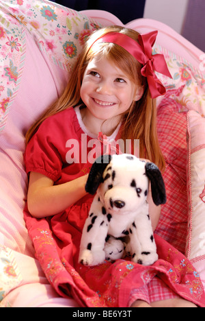 Petite fille portant une robe rouge romantique assis sur son lit et de jouer avec un chien en peluche Banque D'Images