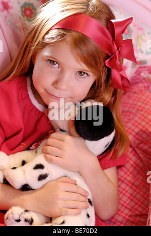 Petite fille portant une robe rouge romantique assis sur son lit et de jouer avec un chien en peluche Banque D'Images