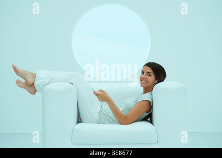 Jeune femme couchée sur un fauteuil, listening to MP3 player, smiling at camera Banque D'Images