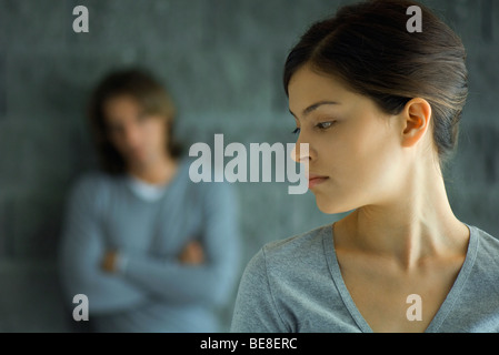 Jeune femme à la recherche sur l'épaule, man in background leaning against wall with arms folded Banque D'Images