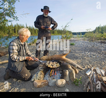 Woman cooking, frire les filets de poisson dans une casserole sur un feu de camp, l'homme avec la plaque en attente derrière, Upper Liard River (Territoire du Yukon, Banque D'Images