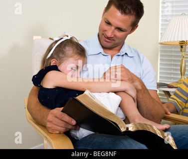 Une petite fille s'endort dans les genoux de son père alors qu'il est en train de lire à ses enfants de la bible. Banque D'Images