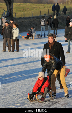 Enfant sur un traîneau poussé par la mère et les promeneurs marchant sur la glace de rivière gelée en hiver Banque D'Images