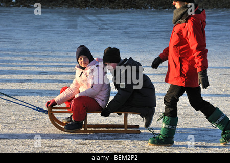 Enfants tiré sur un traîneau et mère à marcher derrière sur la glace de rivière gelée en hiver Banque D'Images