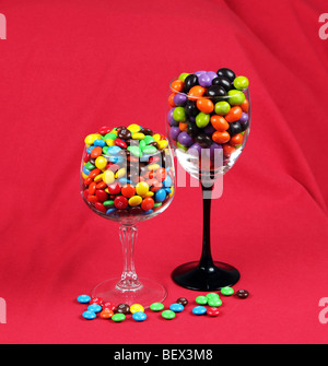 Deux wineglasses de différentes hauteurs rempli de M&Ms placés les uns à côté des autres et tourné sur un fond rouge. Banque D'Images