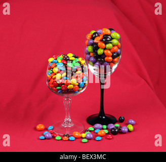 Deux wineglasses de différentes hauteurs rempli de M&Ms placés les uns à côté des autres et tourné sur un fond rouge. Banque D'Images