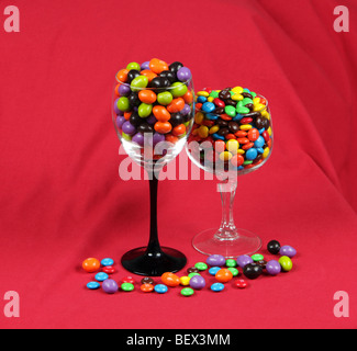 Deux wineglasses de différentes hauteurs rempli de M&Ms placés les uns à côté des autres et tourné sur un fond rouge. Banque D'Images