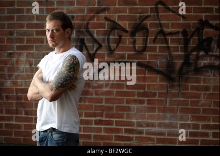 30 ans, homme, et vêtus de t-shirt seul avec les bras tatoués à l'avant du mur de briques couverts de graffitis Banque D'Images