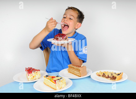 Boy eating cake à une table avec quatre plaques avec d'autres morceaux de gâteau Banque D'Images