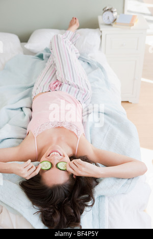 Mixed Race woman plaçant au-dessus des yeux des concombres au lit Banque D'Images