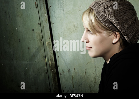 Side portrait d'une jeune femme portant un chapeau tricoté assis devant une vieille porte en acier, de la jeunesse, à l'écart de l'afficheur Banque D'Images