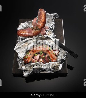 Saucisses grillées et oignons rouges assaisonnés et cuits dans du papier d'aluminium, sur un fond noir. Banque D'Images