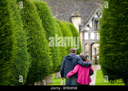 Couple en train de marcher entre les if menant à Lychgate dans cimetière de l'église St Mary Painswick, Angleterre Banque D'Images