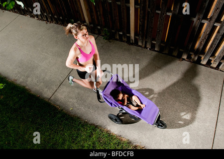 Une edgy-athlétique, à la jeune femme d'un soutien-gorge de sport bénéficie d'une exécution sur un trottoir avec un bébé dans une poussette. Banque D'Images