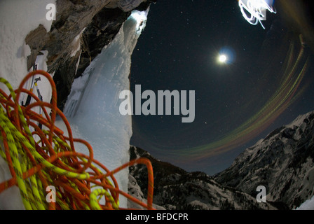 Vue de nuit sur des cordes comme un jeune homme grimpe sur glace avec un projecteur sous une pleine lune. Banque D'Images