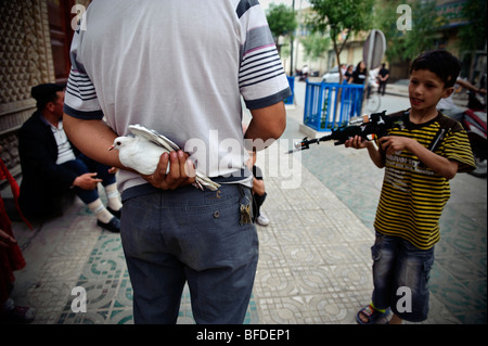 Un homme Ouïghour de la protection d'un pigeon blanc à partir d'un groupe de garçons jouant avec des armes en plastique sur une rue de Kashgar, Xinjiang, Chin Banque D'Images