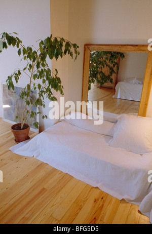 Grand miroir et plante à côté de lit avec draps blanc sur du parquet dans la chambre à coucher appartement moderne Banque D'Images