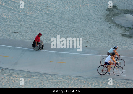 Deux cyclistes passent une personne handicapée exerçant dans un fauteuil roulant sur la même piste cyclable. Banque D'Images