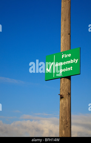 Vert, signe 'Fire Assembly point' sur un poteau télégraphique en bois contre un ciel bleu et nuageux Banque D'Images