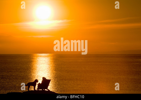 Une femme et son chien sont silhouetté par le coucher du soleil sur un rocher au large d'Oceanside Beach Drive, Victoria BC. Banque D'Images