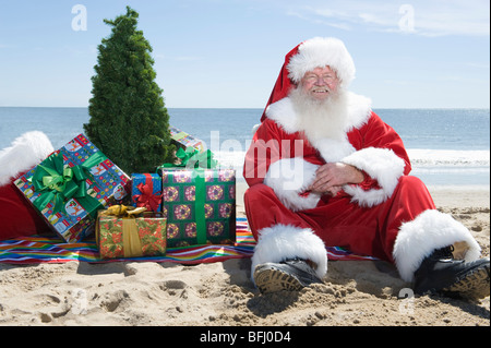 Le Père Noël est assis sur la plage avec un arbre et présente Banque D'Images