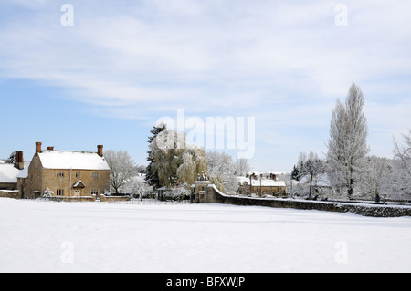 Manor House dans les Cotswolds recouvert de neige avec un village derrière, ciel bleu et nuages. Les peupliers et d'un saule. Banque D'Images