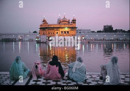 Un groupe de pèlerins s'asseoir et regarder comme le Temple d'or est éclairé au crépuscule. Ce temple, connu comme le Harmandir Sahib, est considéré comme le lieu saint de la religion Sikh. La plupart des sikhs visiter le Temple d'or au moins une fois dans leur vie, en particulier lors d'occasions spéciales comme les anniversaires, mariages, naissances, etc. Le Temple d'Or est situé dans la ville sainte d'Amritsar dans le Pendjab de l'Inde du nord de l'état Banque D'Images