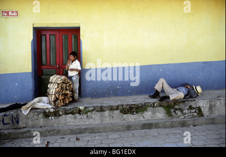 Un homme dort sur le trottoir alors que jeune garçon attend pour charger le bois sur un camion, Chichicastenango, Guatemala Banque D'Images