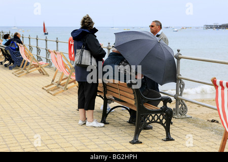 Deux personnes assises sur des sièges au bord de la mer à l'aide d'un parapluie pour l'abri du vent et de la pluie Banque D'Images