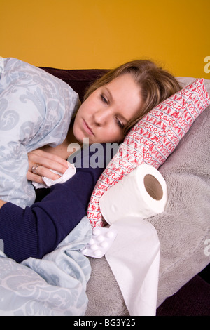 Jeune femme souffrant d'un rhume ou la grippe, recroquevillé sous une couette, avec du papier toilette et des analgésiques à proximité. Banque D'Images