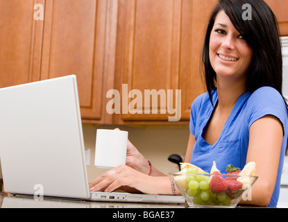 Une jeune femme consulte ses e-mails et travaille sur un ordinateur portable dans la cuisine pendant le petit déjeuner. Banque D'Images