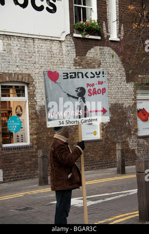 Homme tenant un Banksy pop up shop sign, Covent Garden, London, England, UK Banque D'Images