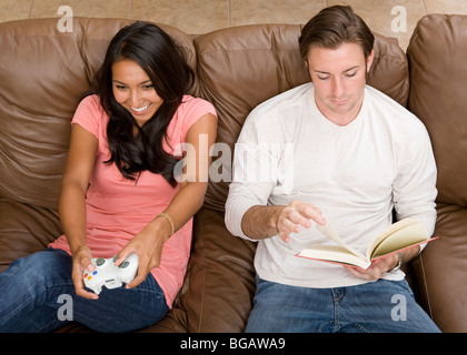 Un jeune couple se détendre à la maison de deux façons différentes ; l'un joue à un jeu vidéo et l'autre lit un livre ensemble sur le canapé. Banque D'Images