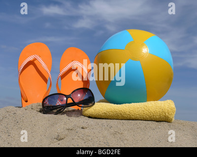 Les tongs Orange conduit à la verticale dans le sable de plage, lunettes, gonflé de ballons de plage et serviette de bain jaune plus de ciel bleu Banque D'Images