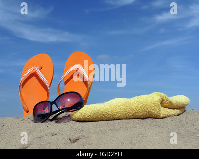Les tongs Orange conduit à la verticale dans le sable de la plage avec des lunettes de soleil et de serviette de bain jaune plus de ciel bleu Banque D'Images