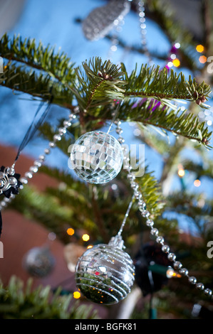 Un mini-miroir boule pend sur un arbre de Noël avec d'autres décorations en décembre dans une baie vitrée. Banque D'Images