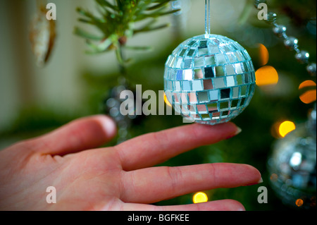 Un mini-miroir balle est main placée sur un arbre de Noël du genre pin dans un salon baie vitrée en décembre. Banque D'Images