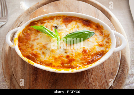 Lasagnes cuites au four avec une croûte de fromage doré garnie d'une feuille de basilic frais, servies dans une casserole blanche sur une planche de bois Banque D'Images