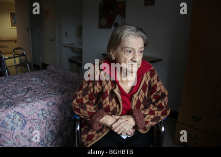 Femme âgée est assis dans sa chambre à une maison de soins infirmiers, perdu dans ses pensées. Banque D'Images