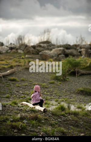 Fille de 5 ans est assis près de rivière, rochers empilés sur une partie de l'horizon de la défense contre les inondations Banque D'Images