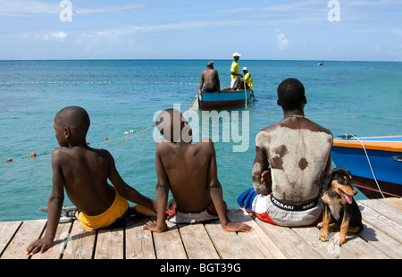 Trois garçons et leur chien assis sur la jetée pour regarder les pêcheurs courriers dans un filet, West Indies. Banque D'Images