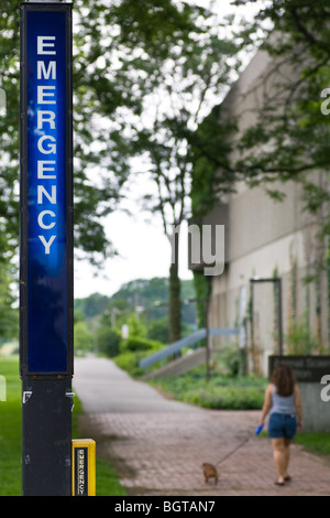 Allumé un pupitre d'appel d'urgence sur un campus universitaire, avec une femme promener son chien le long d'un chemin dans l'arrière-plan. Banque D'Images