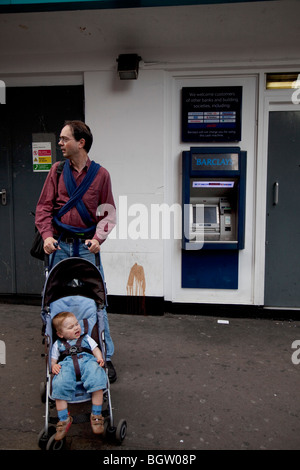 Père avec bébé pousse son enfant dans un landau à l'écart de un distributeur distributeur automatique de billets. Banque D'Images