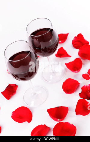 Une paire de verres de vin remplis de vin rouge entourée de pétales de rose rouge sur un fond blanc Banque D'Images