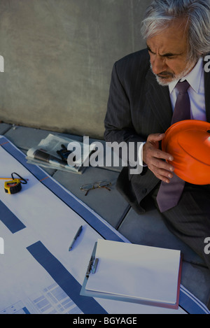 Homme assis sur le sol holding hard hat, looking at blueprints Banque D'Images