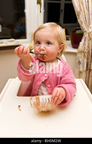 Close up portrait of vertical d'un bébé fille obtenant dans un désordre de manger de la crème glacée au chocolat dans sa chaise haute. Banque D'Images