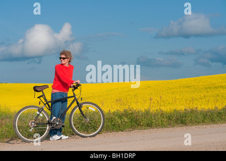 Femme à la mi-50's avec vélo sur route rurale à côté de champ en fleurs de canola, Manitoba, Canada Banque D'Images