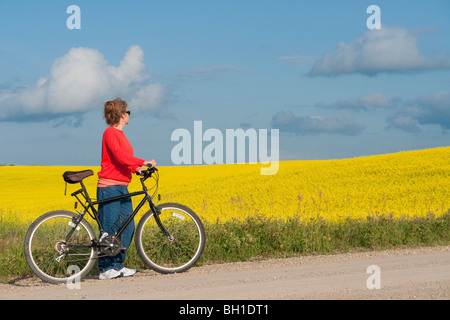 Femme à la mi-50's avec vélo sur route rurale à côté de champ en fleurs de canola, Manitoba, Canada Banque D'Images